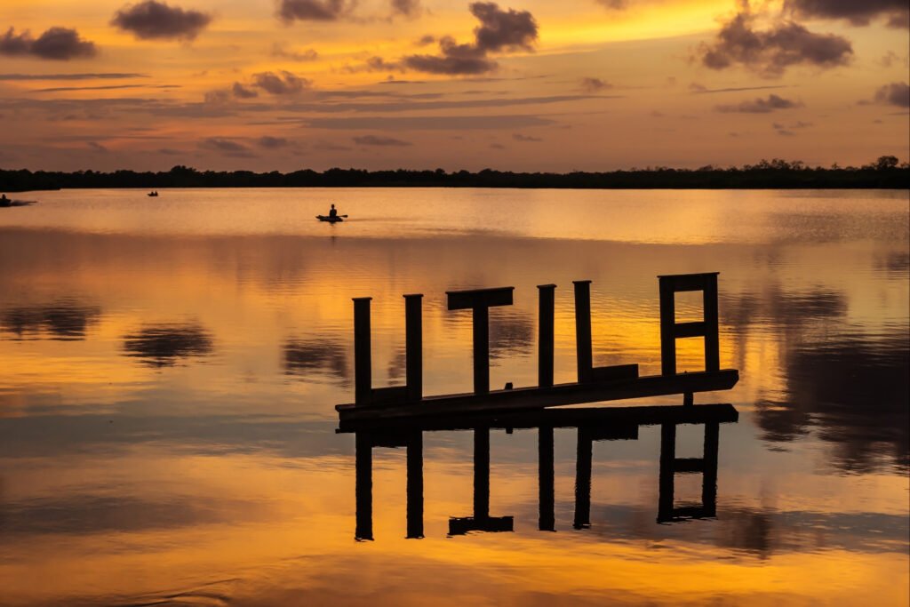 Sunset over the lagoon in Utila, Honduras, with reflective water and wooden "UTILA" sign silhouetted against colorful skies.