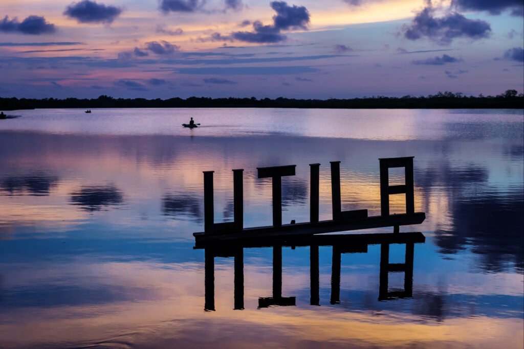 Sunset over the lagoon in Utila, Honduras, with reflective water and wooden "UTILA" sign silhouetted against colorful skies.